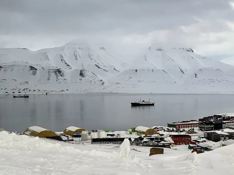 北極圏ロングイェールビーン(Longyearbyen)の港と市街地を丘の上から望む写真。港に浮かぶ船と色とりどりの家並みと背後に雪山が広がる風景